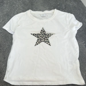 Vit t-shirt med leopardstjärna Lindex - Vit t-shirt från Lindex i storlek 170 med en stor stjärna i leopardmönster på bröstet. Klassisk rund halsringning och korta ärmar. Perfekt för dig som vill ha en basic tee med en cool twist.