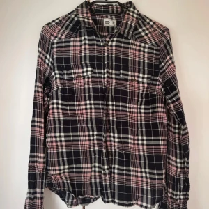 Rutig skjorta i rosa och svart - Säljer en rutig skjorta i svart, rosa och vitt. Skjortan har långa ärmar, klassisk krage och knäppning framtill. Två bröstfickor med knappar. Perfekt för en chill och avslappnad stil.