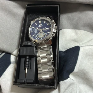 Silverfärgad klocka från SINGDONG - Stilren klocka från SINGDONG med blå urtavla och silverfärgat armband i metall. Klockan har flera detaljerade visare och öppet hjul på urtavlan. Kommer i originalask med verktyg för att justera länken.