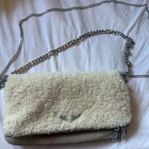 Beige axelväska från Zadig & Voltaire - Superfin beige axelväska från Zadig & Voltaire med fluffig pälsdetalj på locket och cool kedjerem i silver. Väskan har dragkedjor, ett snyggt vingemblem framtill och är gjord i skinn och pälsimitation. Perfekt för dig som vill sticka ut med en unik accessoar.