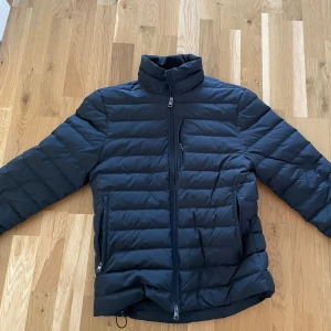 Cos dunjacka - Säljer en svart pufferjacka med hög krage och horisontella sömmar. Jackan har hel dragkedja framtill, två sidofickor med dragkedja och en bröstficka. Perfekt för kyliga dagar och har en clean, stilren look.