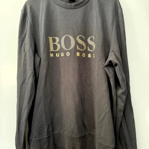 Svart sweatshirt från Hugo Boss - Svart sweatshirt från Hugo Boss med stort gult BOSS-tryck framtill. Tröjan har rund halsringning, långa ärmar och är tillverkad i mjuk bomull. Perfekt för dig som gillar stilrena och klassiska plagg med en tydlig logga.