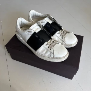 Valentino Garavani sneakers i skinn - Snygga vita sneakers från Valentino Garavani med bred svart rem över mitten och coola nitar baktill. Skorna är i skinn och har klassisk snörning. Perfekta för dig som gillar stilrena men ändå unika detaljer. Använt skick liten skada i sula (se sista bild) original kartong medföljer. Nypris ca 5100kr