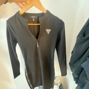 Svart kortklänning från Guess XS - Säljer en svart, figurnära kortklänning från Guess med lång ärm och dragkedja framtill. Klänningen har Guess-logga på bröstet och dekorativt band med logga längs sidan. Materialet är stretchigt och känns mjukt mot huden. Säljs av ingen andvändning.