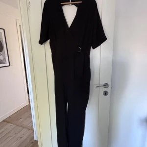 Svart omlott jumpsuit med bälte - Stilren svart jumpsuit med omlott-v-ringning, kort ärm och markerad midja med bälte och silverfärgat spänne. Byxorna är raka och plagget har fickor i sidorna. Snygg detalj med öppen rygg och knäppning i nacken. Perfekt för dig som gillar minimalistisk stil.