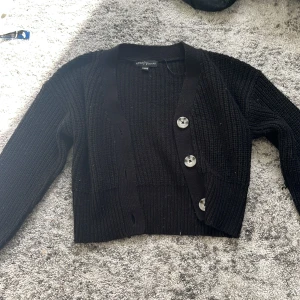 Svart stickad kofta från XOXO Friends - Snygg svart stickad kofta från XOXO Friends med stora silvriga knappar framtill. Modellen är croppad och har långärmad passform. Perfekt att slänga över ett linne för en chill look.