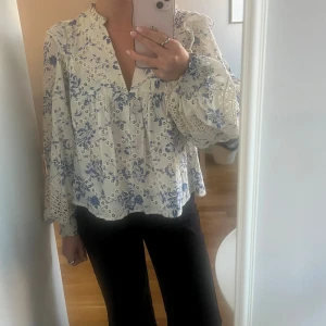 Blommig blus med spetsdetaljer - Romantisk blus i vitt med blått blommönster och v-ringning. Långa ärmar med breda spetsdetaljer och smock vid handlederna. Luftig passform och fina broderade detaljer. Perfekt både till vardags och fest. Använd fåtal gånger, fint skick!