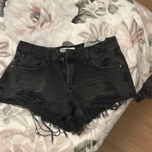 Svarta jeansshorts från Zara - Snygga svarta jeansshorts från Zara med slitna detaljer och fransig kant. Modellen är klassisk femficksdesign med hög midja och råa kanter. Perfekta för sommaren och ger en cool vibe till din outfit.