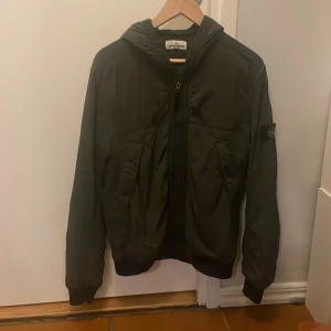 Oliv grön Stone Island vindjacka - Snygg olivgrön Stone Island jacka med huva och patch. Jackan har dragkedja framtill och två stora fickor. Jackan är i bra skick.