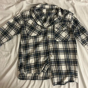 Rutig blå och vit skjorta med knappar - Snygg rutig skjorta i blått, vitt och svart med klassisk krage och två bröstfickor. Skjortan har långa ärmar och stängs med knappar framtill. Perfekt för en avslappnad och cool stil. 