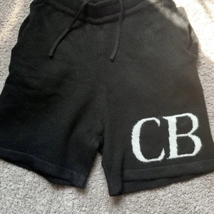 Svarta stickade shorts med CB-tryck - Svarta stickade shorts med vit CB-logga på ena benet. De har snörning i midjan för skön passform och är gjorda i ett mjukt stickat material. Perfekta för chill dagar eller när du vill ha en avslappnad stil.
