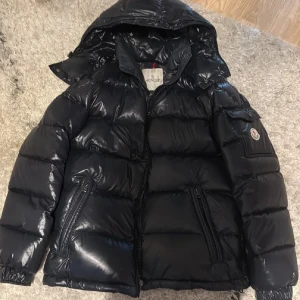 Svart Moncler dunjacka med huva - Pris kan diskuteras vid snabb äffar Säljer en svart, glansig dunjacka från Moncler med huva och dragkedja. Jackan har två snedställda fickor med dragkedja innerficka och en liten ficka med Moncler-logga på ärmen. Insidan har det klassiska serietidningsfodret som är lite av men går att sy tillbaks, Perfekt för kalla dagar. Lappar medföljer 