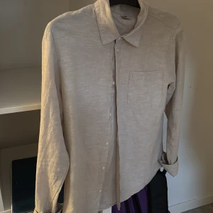 Beige linneskjorta från Lindex - Stilren beige skjorta från Lindex i storlek 170. Skjortan är tillverkad i svalt linne, har klassisk krage, bröstficka och knäppning framtill. Perfekt för dig som gillar en enkel och clean look. Långärmad modell med avslappnad passform.