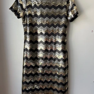 Glittrig kortklänning med paljetter - Säljer en glittrig kortklänning med svart och guldigt sicksackmönster. Klänningen har korta ärmar, rundad urringning på baksidan och är täckt av paljetter som ger en riktigt festlig look. Perfekt för dig som vill sticka ut och glänsa på nästa event!