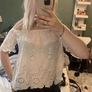 Vit offshoulder blus med broderi - Superfin vit offshoulder blus med broderade detaljer och hålmönster. Blusen har korta ärmar och en lös passform, perfekt för varma dagar. Materialet känns lätt och luftigt, och den har en söt, romantisk vibe med sina blommiga broderier och vågig kant nertill.