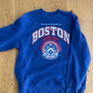 Blå Boston sweatshirt med tryck - Säljer en blå sweatshirt med stort Boston-tryck i vitt och rött på bröstet. Tröjan har rund hals och långa ärmar, samt ett coolt emblem i vitt och blått framtill. Perfekt för dig som gillar college-stil och vill sticka ut med snyggt tryck.