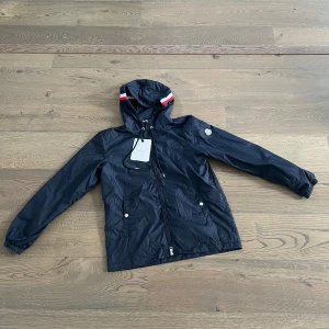 Mörkblå regnjacka från Moncler med huva - Mörkblå regnjacka från Moncler med huva och snygga rödvita detaljer på kanten. Jackan har dragkedja framtill, snörning vid huvan och två fickor med tryckknappar. Perfekt nu till hösten, pris kan diskuteras vid snabbaffär