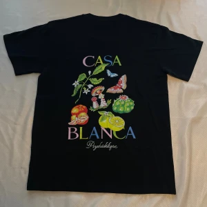 Svart t-shirt från Casablanca M - Svart t-shirt från Casablanca med färgglad print på ryggen med frukter, svampar, fjärilar och blommor samt texten 'Casa Blanca Psychédélique'. Framsidan har en mindre tryckt detalj med svampar och text. Klassisk passform och rund hals. Materialet är mjuk bomull.
