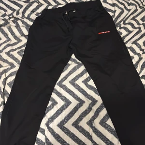 prada byxor - Svarta byxor från Prada med snygg snörning i midjan och diskret röd logga på ena benet. Byxorna har en clean look och är perfekta för dig som gillar stilrena plagg med exklusiv känsla.