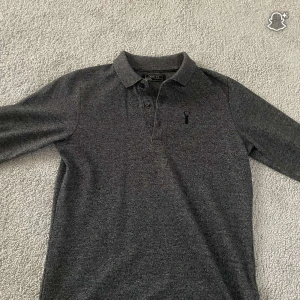 Mörkgrå pikétröja från next - Snygg mörkgrå pikétröja från Next med klassisk krage och tre knappar framtill. Tröjan har ett stilrent broderat motiv på bröstet och är långärmad. Perfekt för dig som gillar enkel och clean stil.