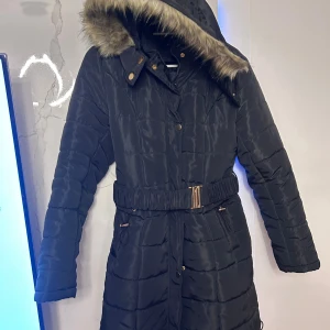 Svart lång pufferjacka Philipp Plein - Snygg svart lång pufferjacka från Philipp Plein med avtagbar huva och fuskpäls. Jackan har guldiga detaljer, bälte i midjan och dragkedjor på fickorna. Perfekt för kalla dagar med sin quiltade design och fluffiga känsla.