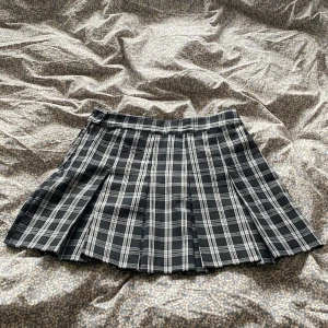 Svartvit rutig plisserad minikjol - Supersnygg svartvit rutig kortkjol med plisserad design och klassisk skoluniform-vibe. Kjolen har en knapp i midjan och är perfekt för dig som gillar en edgy och trendig stil. Passar grymt med boots eller sneakers.