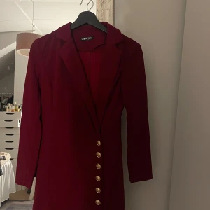 Vinröd kavajklänning  - Elegant vinröd kavajklänning med långa ärmar och v-ringning. Klänningen har guldiga  knappar längs framsidan. 