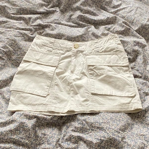 Beige kortkjol med stora fickor - Snygg beige kortkjol med två stora cargofickor framtill och knappstängning i midjan. Kjolen har en rak passform och är tillverkad i ett lätt bomullstyg, perfekt för en chill och avslappnad stil. Passar grymt till sneakers och en enkel t-shirt.