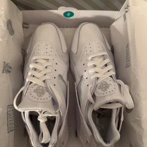 Nike Air Huarache Triple White - Nike Air Huarache i helvitt med klassisk chunky sula och ikonisk hälrem. Ovandel i en mix av skinn och syntet med meshpaneler för extra komfort. Snygg, sportig look med snörning och rund tå. Perfekt för dig som gillar stilrena sneakers med retrovibbar. Helt nya i box!