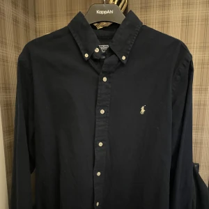 Mörkblå skjorta från Polo Ralph Lauren - Klassisk mörkblå skjorta från Polo Ralph Lauren i custom fit. Skjortan har button down-krage, vita knappar och den ikoniska broderade loggan på bröstet. Tillverkad i mjuk bomull och har lång ärm. Perfekt för en clean och stilren look.