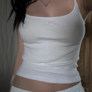 Vit ribbad linnetopp från Brandy - Supersöt vit ribbad linnetopp från Brandy Melville med smala axelband och liten broderad rosa blomma framtill. Toppens material är mjuk bomull 