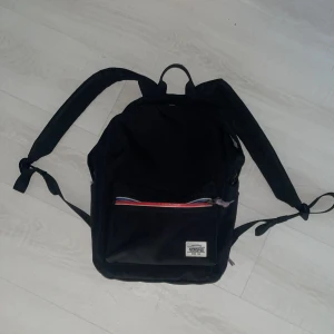 Svart ryggsäck från American Tourister - Snygg svart ryggsäck från American Tourister med klassisk design. Framsidan har en ficka med dragkedja i rött, blått och vitt. Väskan är tillverkad i slitstark polyester och har ett rymligt huvudfack med innerficka och datorfack. Perfekt för skola. 