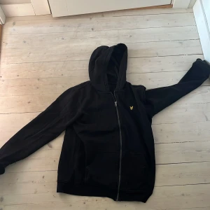 Svart hoodie från Lyle & Scott - Svart hoodie från Lyle & Scott med dragkedja framtill och klassisk huva. Tröjan har en gul broderad logga på bröstet och praktiska fickor. Perfekt för dig som gillar en enkel och stilren look.