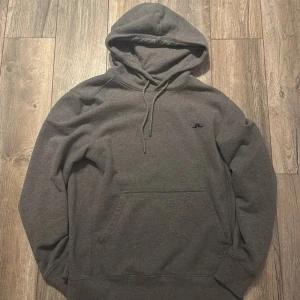 Grå hoodie från J.Lindeberg - Snygg grå hoodie från J.Lindeberg med liten svart logga på bröstet. Klassisk modell med huva och dragsko samt känguruficka framtill. Perfekt för chill dagar och enkel att matcha med jeans eller joggers.