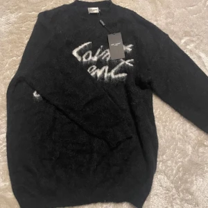 Svart Saint Laurent stickad tröja - Svart stickad tröja från Saint Laurent med vit, handskriven logga framtill. Tröjan har lång ärm, rund hals och en fluffig, lyxig känsla. Perfekt för dig som vill ha en statement piece med exklusiv vibe.