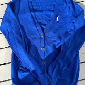 Blå kofta från Ralph Lauren - Säljer en klassisk blå kofta från Ralph Lauren med knappar framtill och broderad logga på bröstet. Koftan har långa ärmar, två fickor och är tillverkad i mjukt bomullsmaterial. Perfekt för lager-på-lager och snygg till jeans eller kjol.