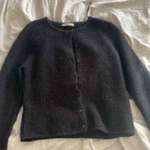 Svart kofta från Gina Tricot - Mysig svart kofta från Gina Tricot.  Klassisk rak modell med rund halsringning och knäppning framtill med svarta knappar. Perfekt för lager-på-lager och passar till allt. Långärmad och fluffig textur som ger en chill vibe. Inte super bra skick eftersom den är väldigt nopprig. 