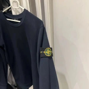 Navy  tröja Stone Island - Svart långärmad tröja från Stone Island med klassisk rund hals och ikonisk logotyp-patch på vänster ärm. Tröjan har en clean och stilren look, perfekt för dig som gillar streetwear och vill ha något som sticker ut lite extra.