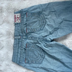 True Religion ljusblå jeans, stl 29 - Säljer ett par ikoniska ljusblå jeans från True Religion med klassiska kontrastsömmar och snygga detaljer på bakfickorna. Bootcut, väldigt slitna därav priset, men det kan fixas om någon är bra på att sy