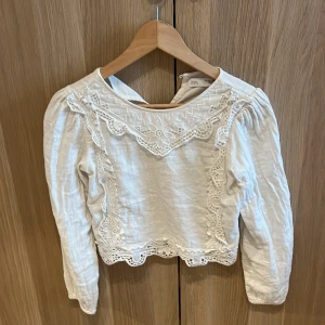 Vit blus med spets från Zara - Superfin vit blus från Zara med långa ärmar och detaljerad spetsbrodyr framtill. Blusen har rund halsringning, broderade blommor och vågig spetskant nertill. Tillverkad i ett lätt och luftigt material som känns skönt mot huden.
