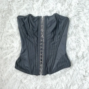 Svart korsett från H&M med hyskknäppning - Säljer en svart korsett från H&M i storlek S. Den har tunna vertikala ränder, hyskknäppning framtill och en snygg, figurnära passform. Storlek S (36), byst 82-86cm, midjan 63-67cm.