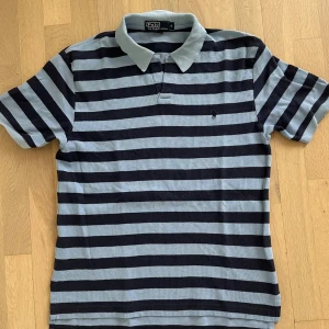 Randig pikétröja från Polo Ralph Lauren - Snygg pikétröja från Polo Ralph Lauren i marinblått och ljusgrått med breda horisontella ränder. Klassisk krage och korta ärmar, perfekt för en chill och stilren look. Broderad logga på bröstet. Gjord i mjuk bomull.