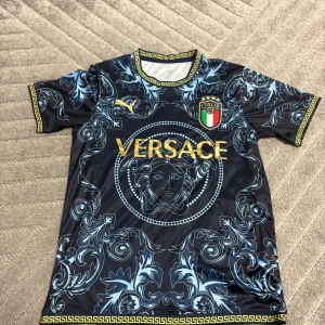 Versace x Puma Italien fotbollströja - Snygg fotbollströja från Puma med Versace-design och Italien-emblem. Tröjan är marinblå med guldiga detaljer och har ett unikt barockinspirerat mönster i ljusblått. Kortärmad modell med gul mönstrad kant vid hals och ärmslut. Materialet är lätt och andas, perfekt för match eller träning.