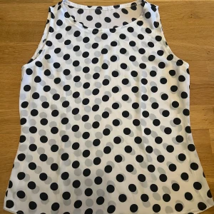 Vit prickig ärmlös linnetopp - Ärmlös linnetopp i vitt med stora svarta prickar. Toppen har rund halsringning och är lätt och luftig, perfekt för varma dagar. Den har en rak passform och är tillverkad i ett mjukt, tunt material som känns skönt mot huden.