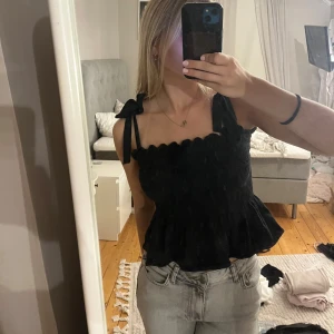 Svart linnetopp med rosettband - Supersöt svart linnetopp med volangkant och knytbara axelband i rosett. Toppen har en croppad passform och smockad överdel som ger en snygg siluett. Perfekt att styla med jeans eller kjol för en trendig look.