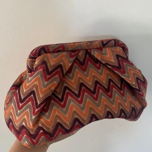 Clutch från bäcksöndergard - Unik clutch från Beck Söndergaard med färgglatt zickzackmönster i orange, rött, beige och vinrött. Väskan är i tyg med innerfoder och har en praktisk innerficka med dragkedja. Inget tecken på användning!