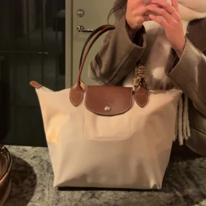 Beige shoppingväska från Longchamp - Säljer en klassisk beige shoppingväska från Longchamp med bruna skinnhandtag. Väskan har en enkel och stilren design med guldknapp framtill och rymligt huvudfack. Perfekt för dig som vill ha en snygg och praktisk väska till vardagen. Går även att vila ihop så den blir liten. Hör av dig om du har några frågor! 🫶🏼💕