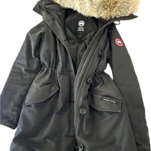 Svart Victoria Parka från Canada Goose med päls - Säljer en svart parkas från Canada Goose med en stor luva med fluffig päls. Jackan har dragkedja, knappar och stora fickor framtill. Perfekt för kalla vinterdagar och riktigt snygg passform. Orginalpris 13 000 kr. Hör av dig vid mer frågor😊