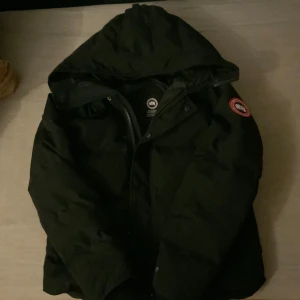 Svart dunjacka från Canada Goose M - Säljer en svart dunjacka från Canada Goose i storlek M. Jackan har huva, dragkedja och klassisk patch på ärmen. Perfekt för kalla dagar med sin puffiga look och stilrena design. Passar dig som vill ha både värme och streetstyle.