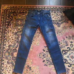 Replay Anbass jeans blå 30/32 - Snygga Replay Anbass jeans i mörkblå tvätt med diskreta slitningar. Modellen har klassisk femficksdesign, smal passform och dragkedja. Perfekt för dig som gillar stilrena och moderna jeans med lite stretch. Midjan är normal och materialet är mjukt jeans. Midja 37 längd 102 (26)